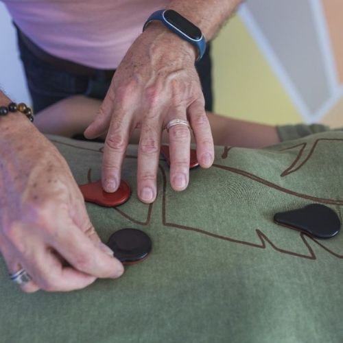 Pessoa deitada, vestindo roupa verde com detalhes em marrom. Sobre ela, as mãos do terapeuta Rui Pereira posicionam ímãs de biomagnetismo.