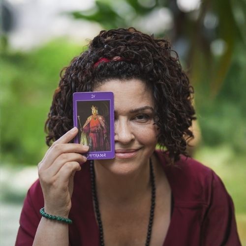 A terapeuta Márcia Hope, de pele clara, usa batom rosa e tem olhos castanho-escuros e cabelos curtos, ondulados e castanho-escuros, presos no alto da cabeça por uma tiara de pano vermelha com detalhes brancos. Em sua mão direita, segura uma carta de tarô à altura dos olhos, cobrindo parcialmente o olho direito. Veste uma blusa na cor vinho, um colar de contas pretas e uma pulseira de contas verdes. Seu rosto expressa um leve sorriso.