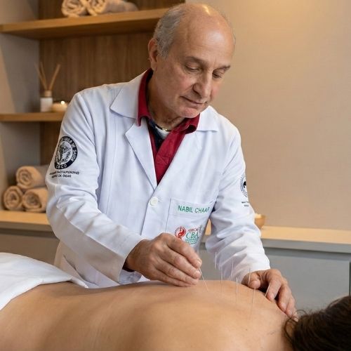 O terapeuta Nabil Chaar, de pele clara, calvo e com cabelos grisalhos curtos nas laterais, veste um jaleco branco, com uma blusa vinho por baixo. Sua mão direita aplica agulhas nas costas de uma mulher, enquanto a mão esquerda serve como apoio ao lado esquerdo das costas. A mulher, de pele clara e cabelos castanho-escuros, está deitada de bruços em uma maca. Suas costas apresentam várias aplicações de agulhas, e ela está coberta por um lençol branco até a altura da cintura.