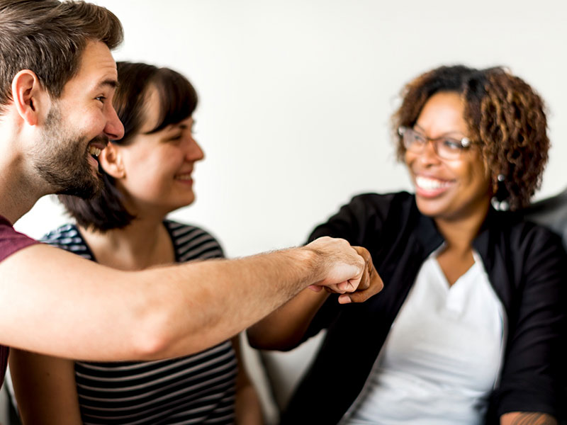 A imagem mostra um momento de interação amigável entre três pessoas em um ambiente interno bem iluminado. No primeiro plano, um homem faz um “soquinho” (fist bump) com outra pessoa, em um gesto de saudação casual que simboliza camaradagem e acordo. À esquerda, um homem jovem com barba sorri enquanto observa a interação. Ao centro, uma mulher com blusa listrada também sorri. À direita, em foco suave, uma mulher negra com cabelos cacheados e óculos sorri abertamente, demonstrando entusiasmo e receptividade. O ambiente transmite positividade, cooperação e informalidade, com fundo desfocado que destaca as expressões e o gesto de conexão entre os participantes.
