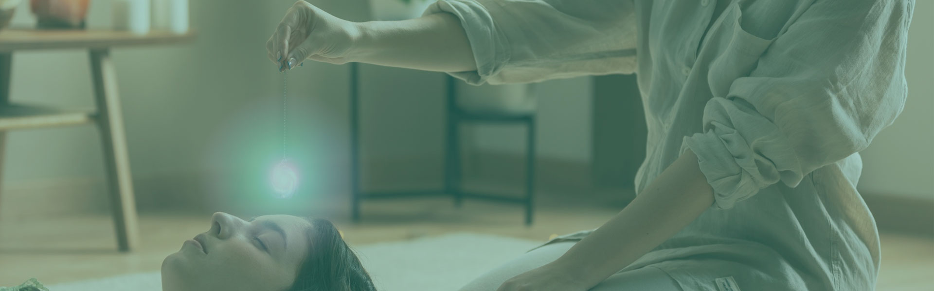 A imagem mostra uma sessão de terapia holística ou trabalho energético, com uma pessoa deitada de costas sobre um colchonete, em estado de relaxamento, e outra pessoa sentada ao lado realizando a prática. A terapeuta mantém as mãos posicionadas acima da cabeça da pessoa deitada, como em uma técnica de imposição de mãos. Um brilho esverdeado ou azulado aparece próximo à cabeça, sugerindo simbolicamente a presença ou foco de energia. O ambiente é interno, com piso de madeira e tapete claro, além de móveis desfocados ao fundo, como mesa e cadeira, criando uma atmosfera calma e serena.