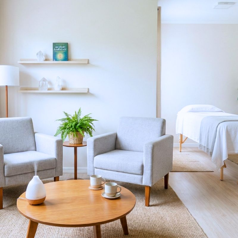 Ambiente acolhedor com uma maca, duas poltronas em tom azul suave e uma mesa redonda com duas xícaras. Há também um difusor ultrassônico liberando “fumacinha”. Uma pequena mesa redonda entre os sofás, com uma samambaia e dois suportes na parede com livros. No canto, um abajur.